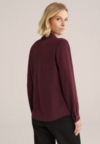 Blusa bordeaux con maniche lunghe, texture morbida e colletto. Presenta un orlo dritto con polsini con bottoni. Indossata sopra pantaloni scuri con texture.
