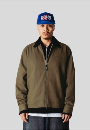 Homme portant une casquette de baseball bleue avec des caractères asiatiques blancs et rouges, une veste zippée vert olive, une couche intérieure noire, une chemise blanche et un pantalon gris.
