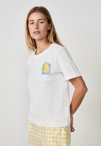 Biała bawełniana koszulka z okrągłym dekoltem, z grafiką cytryny w kolorach niebieskim, żółtym i zielonym oraz napisem "ZEST D'AMOUR". Prosty, klasyczny design.