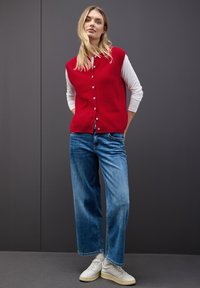 Röd stickad väst med knäppning fram, bärs över en vit långärmad skjorta. Kombineras med blå vida jeans och vita sneakers.