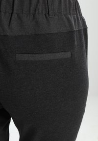 Calças pretas com um cós elástico, apresentando uma textura suave e um bolso traseiro com uma abertura horizontal para fácil acesso.