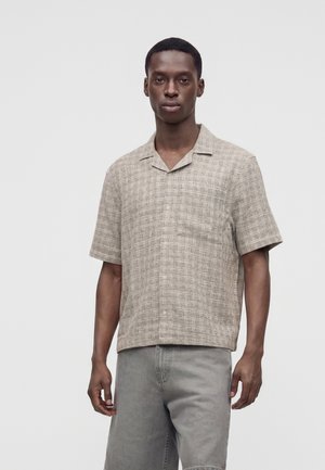 Man draagt een beige geruit overhemd met korte mouwen en kraag en een grijze broek, staand tegen een effen lichte achtergrond.