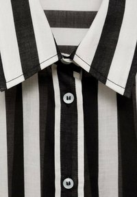 Camicia a righe bianche e nere con colletto a punta, chiusura con un singolo bottone e un tessuto leggero dall'aspetto leggermente texture.