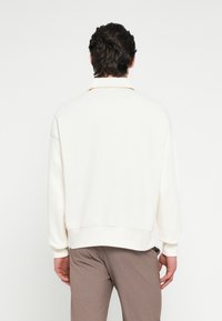 Felpa color crema con vestibilità rilassata, polsini a coste e un design del colletto sottile. Materiale morbido con uno stile minimalista.