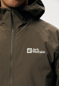 Brun vandtæt jakke med høj krave og lynlås, der har et hvidt "Jack Wolfskin" logo. Materialet ser glat og let ud.