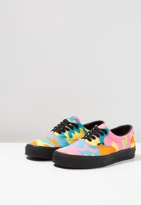 Baskets basses avec semelles et lacets noirs, présentant un motif camouflage coloré en rose, bleu, jaune et orange.