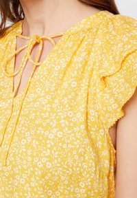 Blusa floreale gialla con una texture morbida, maniche arricciate e chiusura con nodo anteriore, decorata con motivi floreali bianchi.