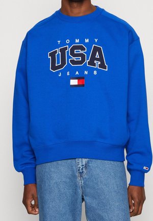 Niebieska bluza z napisem „TOMMY USA JEANS” i logo flagi Tommy Hilfiger, noszona z jasnoniebieskimi jeansami.