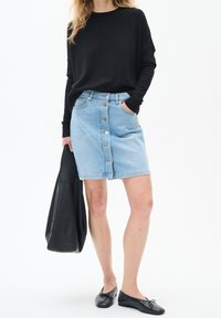 Kvinna i en svart långärmad topp, ljusblå knäppt denimkjol, svarta ballerina skor och håller en stor svart tote väska.