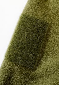 Gros plan d'un tissu polaire vert montrant un patch rectangulaire de boucle texturée cousu sur la surface.