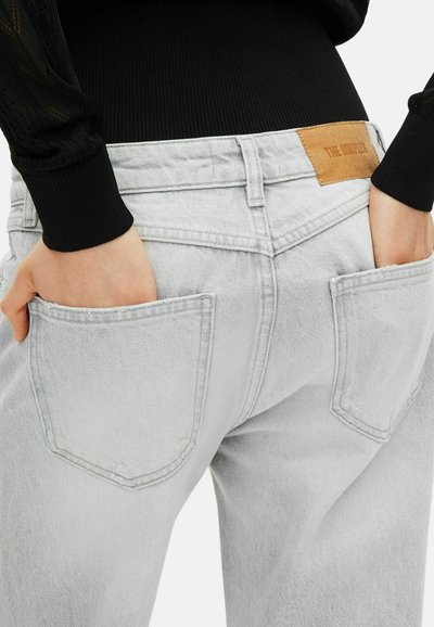 Manos en los bolsillos traseros de unos jeans de mezclilla gris claro, combinados con una camiseta de manga larga negra de canalé, parche de cuero visible en la cinturilla.