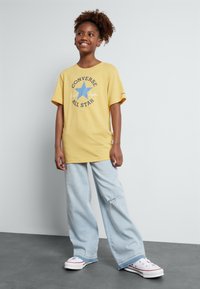 Gul bomulds t-shirt med "Converse All Star" grafik, der har en blå stjerne, parret med lyseblå vidde ben jeans og hvide sneakers.