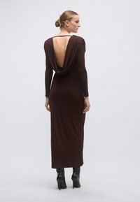 Μακρύ, βαθύ καφέ φόρεμα με ντραπαρισμένη πλάτη, μακριά μανίκια και εφαρμοστή σιλουέτα. Μαύρες, γυαλιστερές μπότες με ψηλό τακούνι ολοκληρώνουν το ντύσιμο.