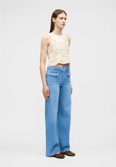 Νεαρή γυναίκα στέκεται φορώντας αμάνικο κρεμ κορμάκι από βελονάκι, μπλε τζιν παντελόνι με φαρδιές ψηλές μέσες και επίπεδα παπούτσια με λεοπάρ μπορντούρα.