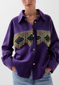 Camicia in suede viola con un design a frange di perline, arricchita con accenti verdi e rossi, dettagli in oro e maniche lunghe. Vestibilità casual.