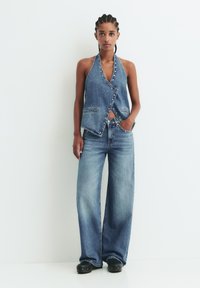 Denim halterneck trikotnica s zavitim dizajnom in gumbi, v kombinaciji s širokimi denim hlačami. Obe v svetlom modrem pranju. Gladka tekstura.