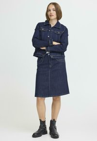 Mörkblå denimjacka med knappar, kombinerad med en knälång denimkjol, kompletterad med svarta ankelstövlar, mot en neutral bakgrund.