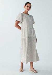 Robe en coton rayée jusqu'aux genoux, avec des manches courtes bouffantes, un col rond et une jupe à volants. Présente une couleur beige clair avec de fines rayures brunes.