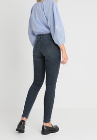 Jeans ajustados de color azul oscuro con una cintura de media altura, hechos de denim elástico. Con bolsillos traseros sencillos y un acabado liso. Combinados con una blusa a rayas.