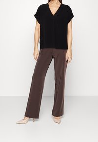 Femme portant un blouse noir à manches courtes en V, un pantalon large marron et des escarpins nude à bout pointu, se tenant devant un fond blanc.