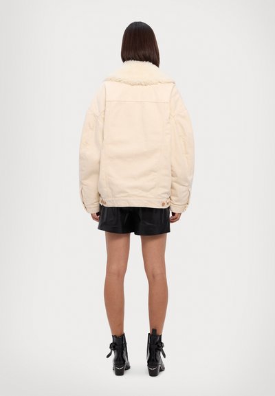 Chaqueta de gran tamaño color crema con cuello de borreguito, con mangas amplias y detalles de botones, combinada con pantalones cortos negros y botas de tobillo.