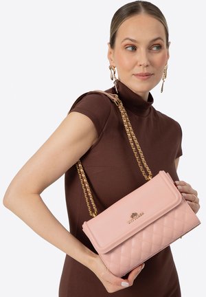 Mujer con vestido marrón sosteniendo un bolso acolchado de color rosa claro con correas de cadena dorada sobre el hombro, luciendo pendientes dorados de diseño escultórico.