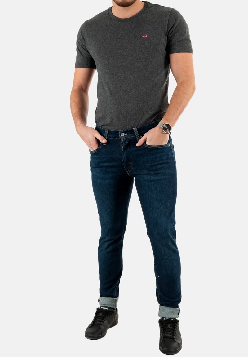 Graues Baumwoll-T-Shirt mit kleinem rotem Logo, kombiniert mit dunkelblauen Slim-Fit-Jeans und schwarzen Sneakers, die mit hochgerollten Ärmeln präsentiert werden.