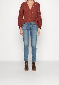 Rostfarbenes Wickeltop mit langen Puffärmeln und floralen Stickereien, kombiniert mit engen blauen Jeans und braunen Absatzstiefeln.