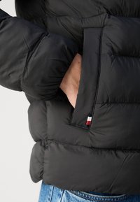 Juodas pūkinis švarkas su siuvinėtu paviršiumi, turintis šoninį kišenę ir mažą vėliavos akcentą raudonos, baltos ir mėlynos spalvų.