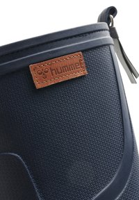 Botte en caoutchouc bleu marine avec surface texturée et une étiquette en cuir marron portant un logo embossé. Comprend une languette pour un enfilage facile.