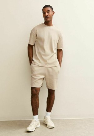 Man staat tegen een beige muur en draagt een beige shirt met korte mouwen, bijpassende korte broek, witte sokken en crèmekleurige sneakers.