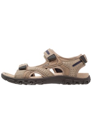 Beige sandal med perforeret ruskindslignende overdel, justerbare remme med velcro, og en robust sort gummisål.