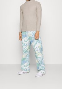 Person klädd i ett beige polokragsweater, pastellgröna och blå marmorering vid vid byxor, samt vita sneakers som står mot en enfärgad bakgrund.