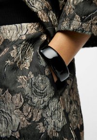 Bague brillante noire sur une main, contrastant avec le tissu texturé noir et doré à motif floral de l'habillement.