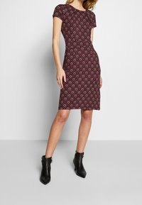 Abito aderente a maniche corte con una base bordeaux e motivi floreali geometrici in crema e verde; lunghezza fino al ginocchio. Viene indossato con stivali neri alla caviglia.