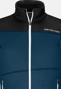 Blaue und schwarze Zip-Jacke mit hohem Kragen, strukturiertem Stoff, vertikalen Nahtdetails und weißem Reißverschluss. "ORTOVOX"-Logo auf der Brust.