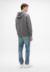 Hanorac gri cu glugă și șnur, purtat împreună cu blugi dărâmați de culoare albastră deschisă și adidași albi, văzut din spate. Textură netedă.