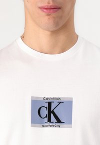 White cotton t-shirt featuring a rectangular graphic in blue and black with "Calvin Klein," "CK," and "New York City" text.