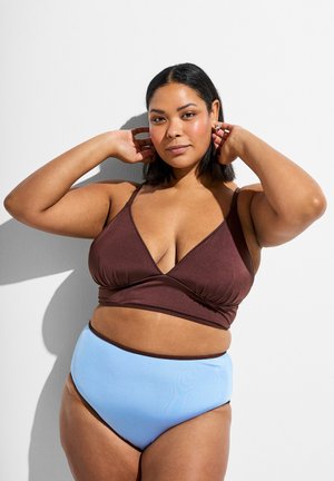 Naine poseerib käed peaaegu pea juures, kandes pruunikat bralette'i ja kõrge vöökohaga hele sinist bikiini alumist osa valgel taustal.