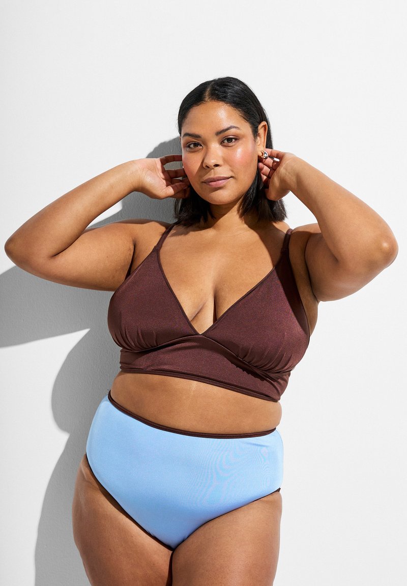 Kvinde poserende med hænderne nær hovedet iført en brun bralette og lyseblå bikinitrusser med høj talje mod en hvid baggrund.