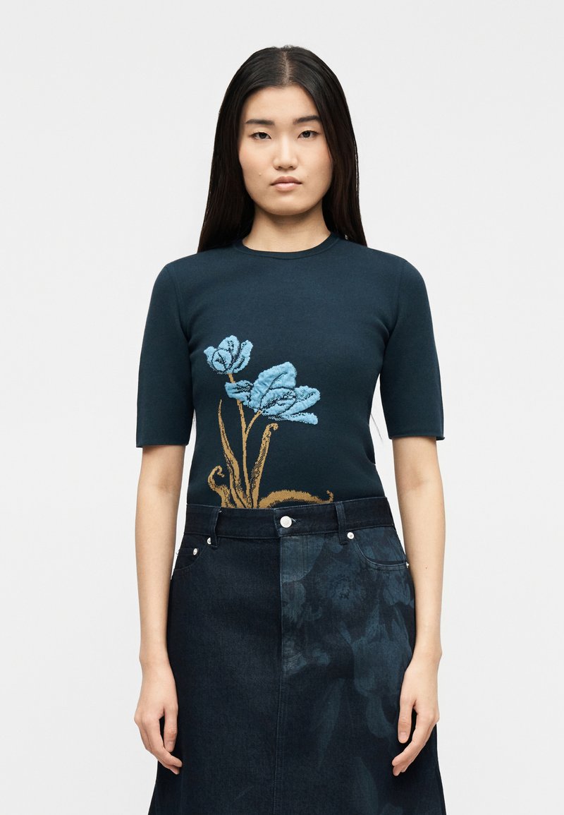 Γυναίκα με μακριά μαλλιά φοράει μπλε σκούρο κοντομάνικο τοπ με υφασμένα ανοιχτό μπλε λουλούδια και μπλε σκούρη τζιν φούστα με διακριτικό floral σχέδιο.