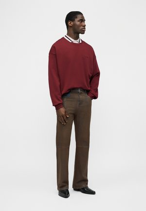 Moški stoji, oblečen v bordo majico s kapuco preko bele srajce s ovratnikom, rjave hlače in črne usnjene čevlje na enostavni belem ozadju.