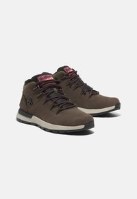 Botas de senderismo de ante marrón de media altura con acentos de malla negra, cordones morados y suela de goma. Presenta un logo en el lateral y un collar acolchado.