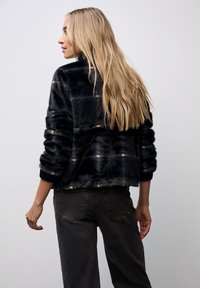 Sort faux fur-jakke med strukturerede bånd i lysere nuancer. Har en høj krave og ribbede manchetter. Sammen med mørkegrå bukser.