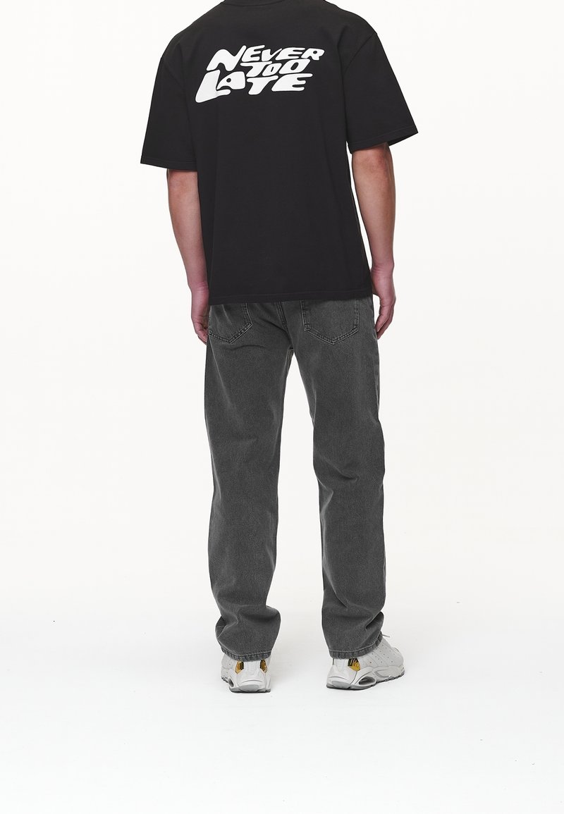 Camiseta negra con el texto blanco "NUNCA ES TARDE" en la parte de atrás, combinada con jeans grises y zapatillas blancas con acentos amarillos.