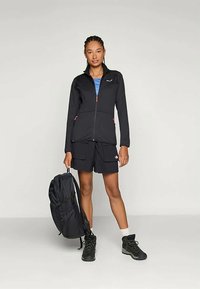 Kvinne står iført svart zip-up jakke, svarte cargo-shorts, tursko, holder en svart ryggsekk ved siden av mot en ensfarget bakgrunn.