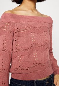 Różowy sweter dzianinowy z odkrytymi ramionami, charakteryzujący się textured wzorem fal oraz ażurowymi detalami na ciele i rękawach.