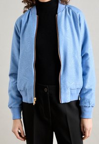 Chaqueta bomber de color azul claro con textura de paisley, cremallera dorada y puños acanalados. Llevada sobre un cuello de tortuga negro, combinada con pantalones negros.