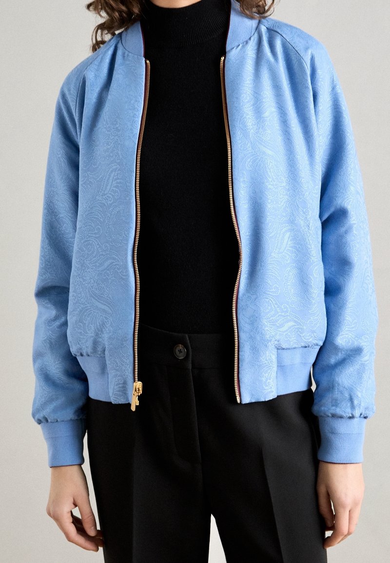 Chaqueta bomber de color azul claro con textura de paisley, cremallera dorada y puños acanalados. Llevada sobre un cuello de tortuga negro, combinada con pantalones negros.