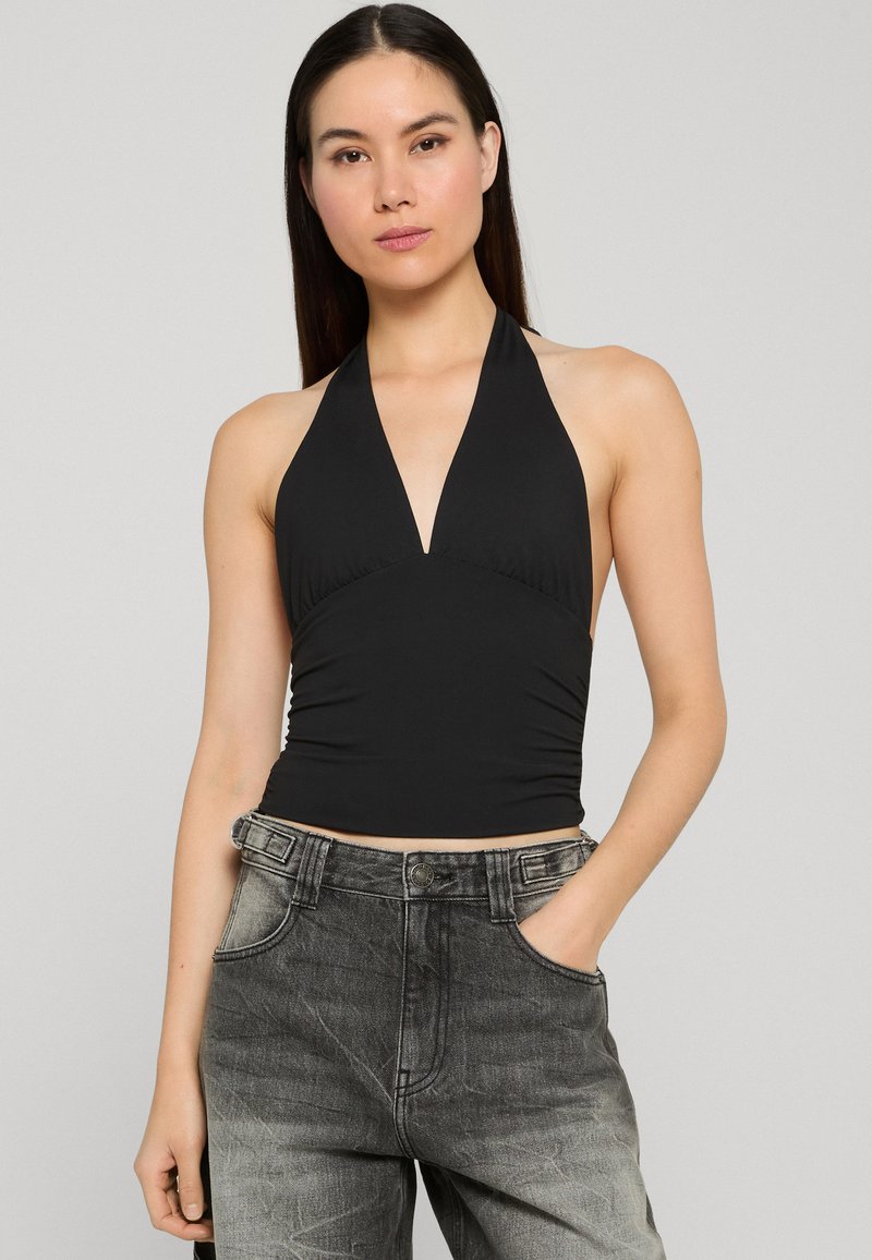 Femme aux longs cheveux bruns portant un débardeur noir et un jean gris délavé taille haute, debout avec une main dans la poche.